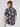 A young man with light brown hair faces away in a BENJAMIN-BLACK navy palm tree print 4-way stretch shirt by Projek Raw and white pants, standing against a plain light background.