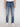 A person wears Projek Raw GABRIEL-DISTRESSED INDIGO jeans, paired with white sneakers and a white shirt, standing against a plain light background. Only the lower torso and legs are visible.