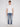 A young person with short light brown hair stands facing forward against a plain light background, wearing a white t-shirt, Projek Raw XANDER-DISTRESSED INDIGO jeans, and white sneakers.