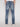 A person wearing a white shirt and Projek Raw XANDER-DISTRESSED INDIGO jeans stands with their back to the camera. The jeans have back pockets, a waistband leather patch, and are styled with white sneakers.