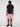 A man stands facing slightly left, wearing the Projek Raw NATHANIAL-ROSE black short-sleeve button-up shirt, pink knee-length shorts, and black sneakers against a plain white background.