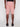 A person wears Projek Raw’s NATHANIAL-ROSE light pink knee-length shorts with visible back pockets and stitching, paired with black shoes, and stands facing away from the camera against a plain white background.