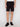 A person wearing black CYPRIAN-BLACK drawstring shorts by Projek Raw, paired with a white shirt and black sneakers, stands against a plain light background. The image is cropped from shoulders to feet.