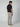 A man faces away, glancing over his shoulder. He wears a black short-sleeve polo, PROJEK RAW Mens 5-Pocket Solid Bungee Pant in light beige, and beige loafers against a plain light background.