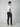 A man stands turned slightly to the side, wearing a white t-shirt, PROJEK RAW Mens Bungee Chino Pant in dark tones, and beige slip-on shoes against a light grey background, glancing over his shoulder with effortless casual style.