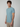 A young man with curly brown hair wears a light blue PROJEK RAW Mens Short Sleeve Crew Neck T-shirt and beige pants, standing against a plain light gray background, looking at the camera with a neutral expression.