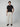 A young man stands against a plain gray background, wearing PROJEK RAWs Mens Textured Bungee Melange Chino Pant in light beige, paired with a black t-shirt and brown loafers. His left hand is in his pocket as he faces the camera with a neutral expression.