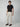 A man stands against a plain background wearing a black short-sleeve t-shirt, brown loafers, and PROJEK RAW Mens Textured Bungee Melange Chino Pant in light beige. He looks slightly to the side with a neutral expression.