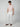A man stands against a plain background wearing a light pink t-shirt, PROJEK RAW Mens Drawstring Linen Pant in white, and brown sandals. His hands are in his pockets and he is looking downwards.