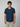 A young man with curly brown hair models the PROJEK RAW Mens Short Sleeve Textured 4-Way Stretch Shirt in dark blue with beige pants, standing against a plain light gray background.
