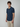 A young man with short curly hair and a left forearm tattoo stands against a plain background, wearing PROJEK RAW’s Mens Short Sleeve Textured 4-Way Stretch Shirt in dark blue with a patterned collar, paired with light beige pants.