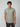 A young man with curly brown hair wears the PROJEK RAW Mens Short Sleeve Textured 4-Way Stretch Shirt in light gray with dark pants, standing facing forward with one hand in his pocket against a plain, light background.