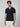 A man with curly brown hair wears a PROJEK RAW Mens Short Sleeve Knit Polo Sweater in navy with white trim and a chest pocket, paired with white pants, standing against a plain light gray background.