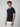 A man with curly brown hair wears a PROJEK RAW Mens Short Sleeve Knit Polo Sweater in dark blue with white trim and buttons, paired with white pants, standing against a plain light gray background.