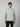 A young man with straight brown hair stands facing forward, wearing a light gray PROJEK RAW Mens Short Sleeve Crew Neck T-shirt and black pants, ideal for casual wear, set against a plain light-colored background.