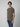 A young man with brown hair stands sideways, wearing a PROJEK RAW Mens Short Sleeve Carbon Peached Pigment Wash Jersey T-shirt in olive green and blue jeans against a light gray background for a casual vintage look.