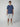 A young man stands facing forward against a plain light gray background, wearing a PROJEK RAW Mens Short Sleeve Graphic T-Shirt with a chest pocket, paired with light gray knee-length shorts and white sneakers.
