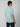 A young man with brown hair is seen from behind, wearing a PROJEK RAW Mens Short Sleeve Carbon Peached Pigment Wash Jersey T-shirt in light blue and dark pants, standing against a plain light gray background.