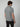 A person with medium-length brown hair is standing and facing away from the camera, wearing a PROJEK RAW Mens Short Sleeve Marled Jersey Polo in light gray and black pants against a plain light background.