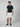 A man stands facing forward in a navy short-sleeve polo, PROJEK RAW Mens 5-Pocket Solid Bungee Shorts in white, a dark belt, and navy slip-on shoes against a plain light gray background.
