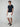 A man faces forward with a relaxed expression, wearing PROJEK RAW Mens 5-Pocket Solid Bungee Shorts, a navy short-sleeve polo, belt, and navy slip-on shoes against a plain background.
