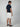 A man stands facing away, wearing a navy short-sleeve polo, PROJEK RAW Mens 5-Pocket Solid Bungee Shorts, and navy slip-on shoes. The background is plain and light-colored.