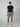 A young man stands against a plain light gray background, wearing a black t-shirt, PROJEK RAW STRIPE BUNGEE CHINO SHORT with a striped pattern made from stretch fabric, and white sneakers. He faces the camera with a neutral expression and relaxed posture.