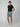A young man stands against a plain background wearing a black t-shirt, PROJEK RAW STRIPE BUNGEE CHINO SHORT, and white sneakers, one hand in his pocket and a neutral expression.