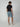 A man stands facing away, wearing a black T-shirt, PROJEK RAW STRIPE BUNGEE CHINO SHORT in light blue with stretch fabric, and white sneakers. The background is plain and light-colored.