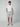A man faces forward against a light gray background, wearing a plain white t-shirt, PROJEK RAW MENS CHINO SHORT in tailored light gray, and gray sneakers, with one hand in his pocket.