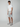 A man with short curly hair and a neutral expression wears a white t-shirt, PROJEK RAW MENS CHINO SHORT in light gray (tailored fit), and gray and white sneakers, standing against a plain light background.