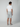 A man faces away from the camera wearing a white short-sleeve shirt and light gray PROJEK RAW MENS CHINO SHORT, featuring a tailored fit and breathable fabric, paired with gray sneakers against a plain light background.