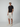 A man stands in a studio wearing a black T-shirt, PROJEK RAW MENS CHINO SHORT, beige sneakers, and a beige cap. With one hand in his pocket, he looks towards the camera against a plain light background.