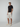 A man stands against a plain light background, wearing a black T-shirt, PROJEK RAW MENS CHINO SHORT made of breathable fabric, cream sneakers, and a beige baseball cap. He faces the camera with a neutral expression and has one hand in his pocket.