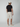 A man faces away, wearing a black t-shirt, PROJEK RAW MENS CHINO SHORT in light beige with a tailored fit, white sneakers, and a white cap against a plain light gray background.