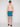 A young man faces the camera with a neutral expression, wearing Projek Raw’s BRUNO-TURQUOISE swim trunks featuring a palm tree pattern, along with sunglasses and black slide sandals. He is shirtless against a plain background.