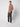 A man stands in a studio against a light gray background, wearing PROJEK RAW Men’s Textured Bungee Pull-on Chino Pant in dark gray, a brown half-zip sweater, and white sneakers, with his left hand in his pocket and facing slightly to the side.