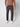 A person wearing a brown long-sleeve shirt, white sneakers, and PROJEK RAW Men’s Textured Bungee Pull-on Chino Pant in dark gray stands against a plain light background with one hand in their pocket.