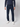 A person models PROJEK RAW Mens 5-Pocket Flex Knit Jeans in dark blue with a navy sweater and white sneakers, standing against a light gray background. The head is not visible in the image.