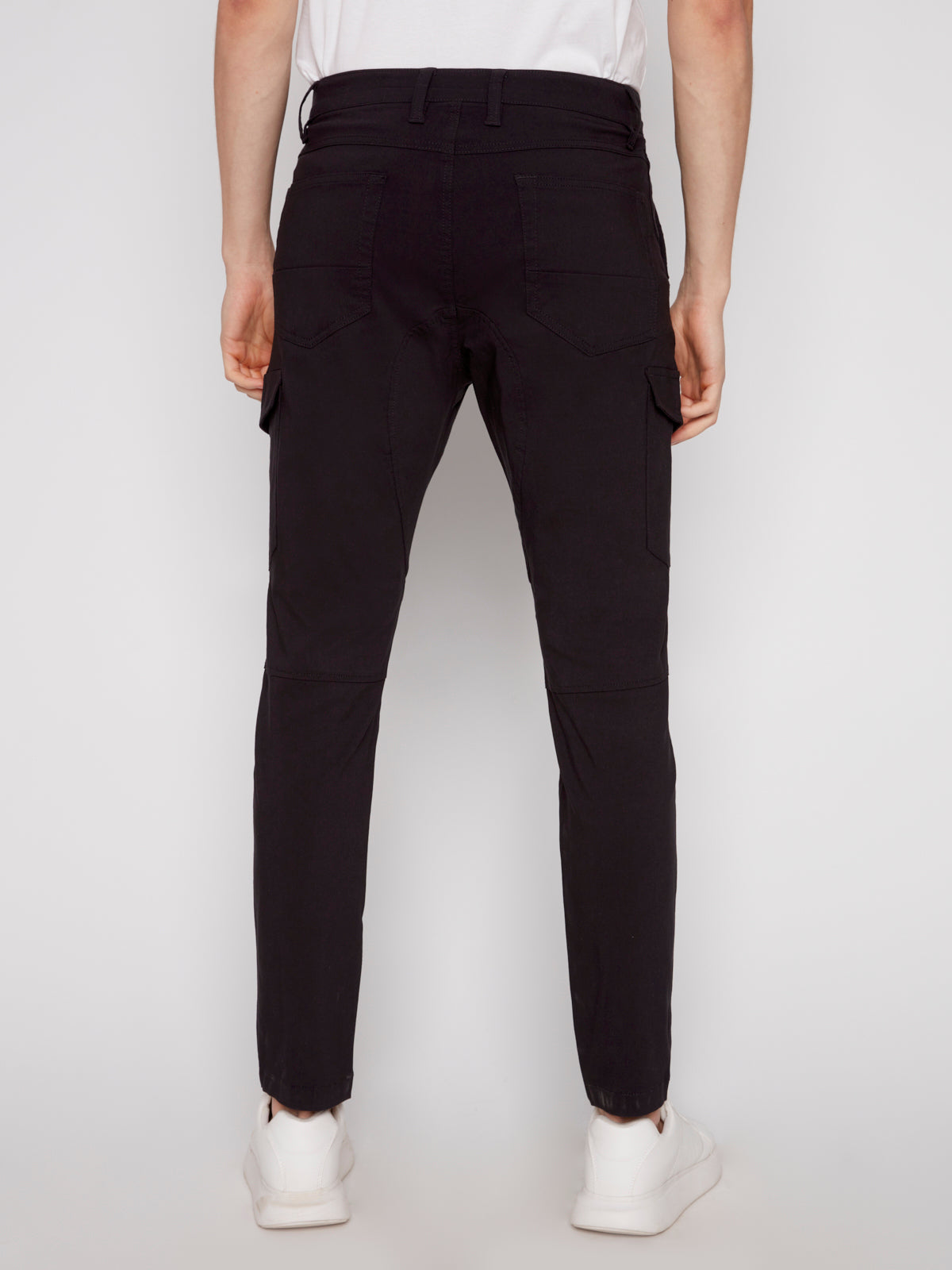 A person is shown from the back in STEPHANE-BLACK cargo pants by Projek Raw, made from bungee fabric, featuring visible back and side cargo pockets, paired with white sneakers against a plain light background.