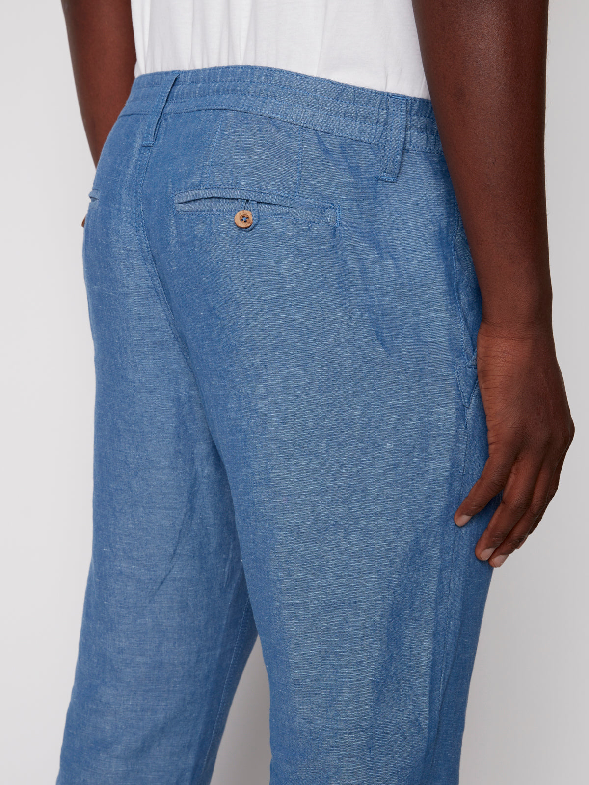 A close-up of a person wearing Projek Raws LUC-BLUE light blue linen drawstring pants with an elastic waistband and back pocket, paired with a white shirt for a relaxed vacation-ready look.