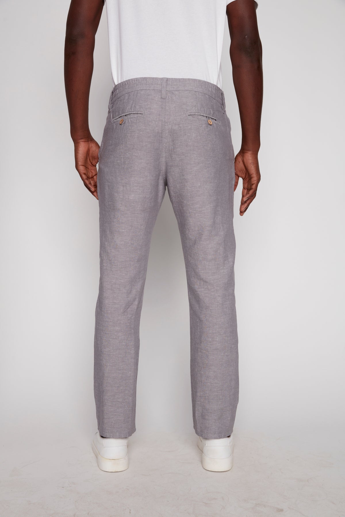 Dressed in Projek Raw’s LUC-GREY linen drawstring pants and a white tee, a person stands with their back to the camera against a plain white background, highlighting an effortlessly relaxed summer look.