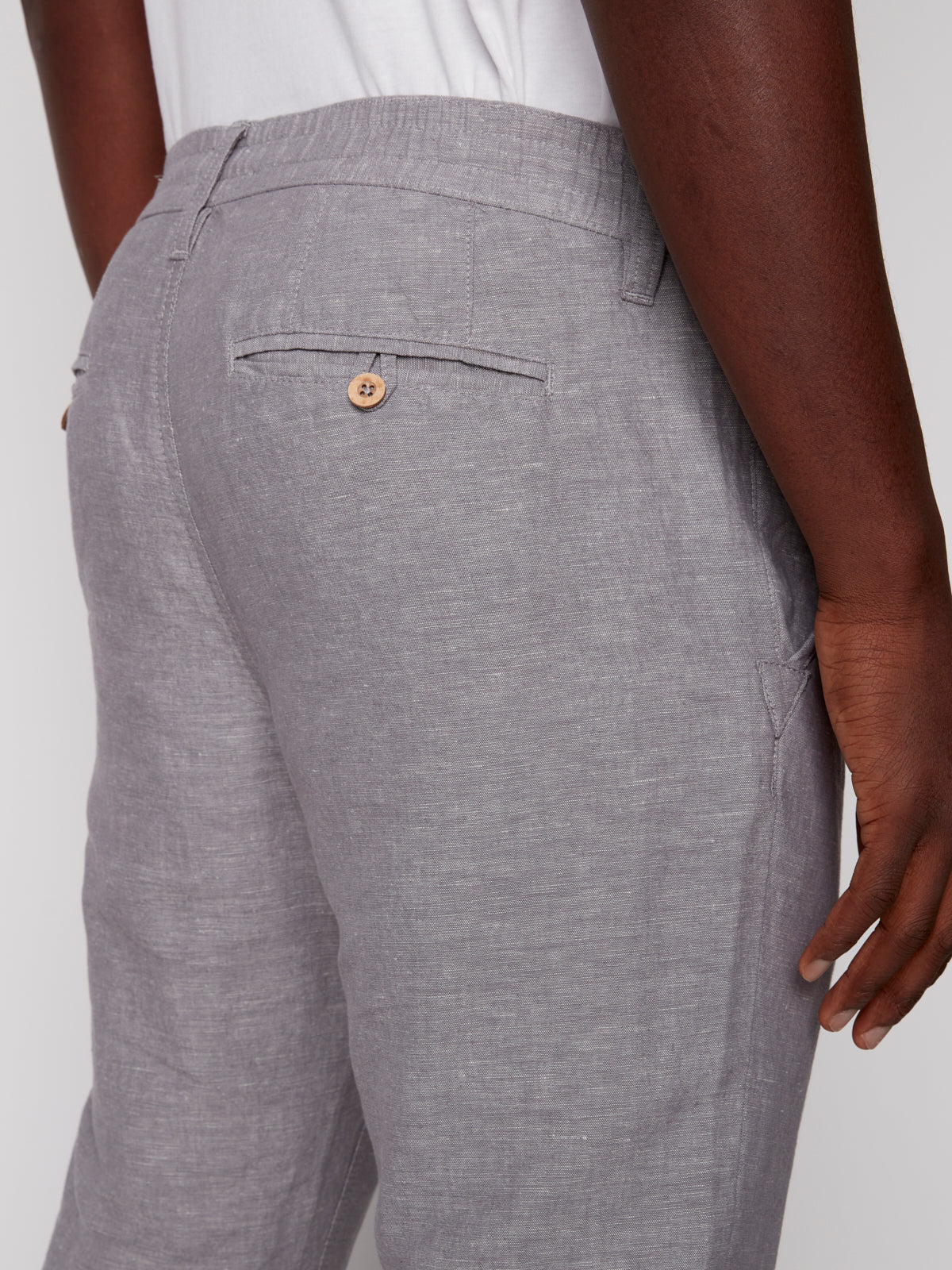 A person shown from behind at waist level wears Projek Raws LUC-GREY light gray linen drawstring pants and a white shirt, with one hand by their side and a buttoned back pocket—a relaxed summer style.