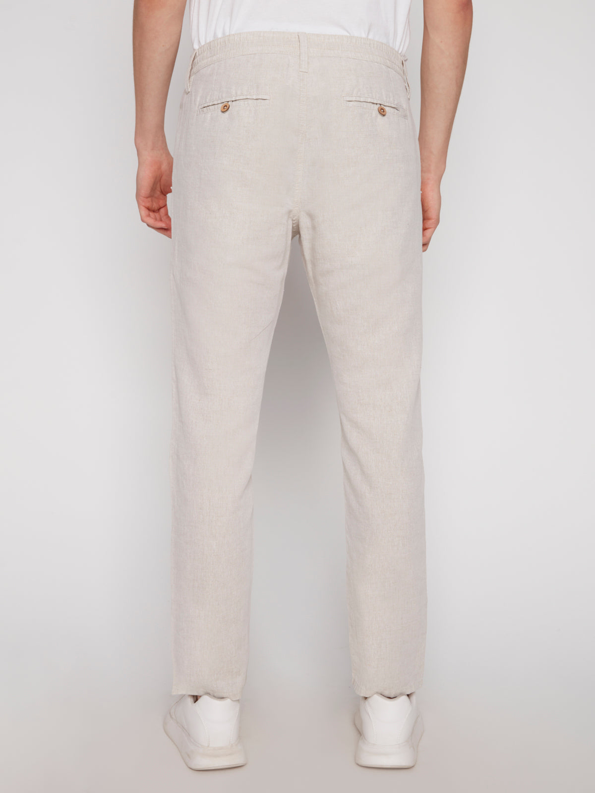 A person is shown from the back wearing Projek Raw’s LUC-NATURAL vacation-ready linen drawstring pants in light beige, paired with white sneakers. The summer pants feature two back pockets with button closures, set against a plain white background.