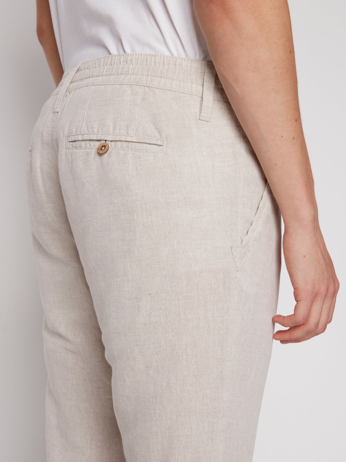 A person is seen from the side wearing Projek Raws LUC-NATURAL linen drawstring pants and a white shirt, highlighting the elastic waistband, detailed seams, and back pocket with button.