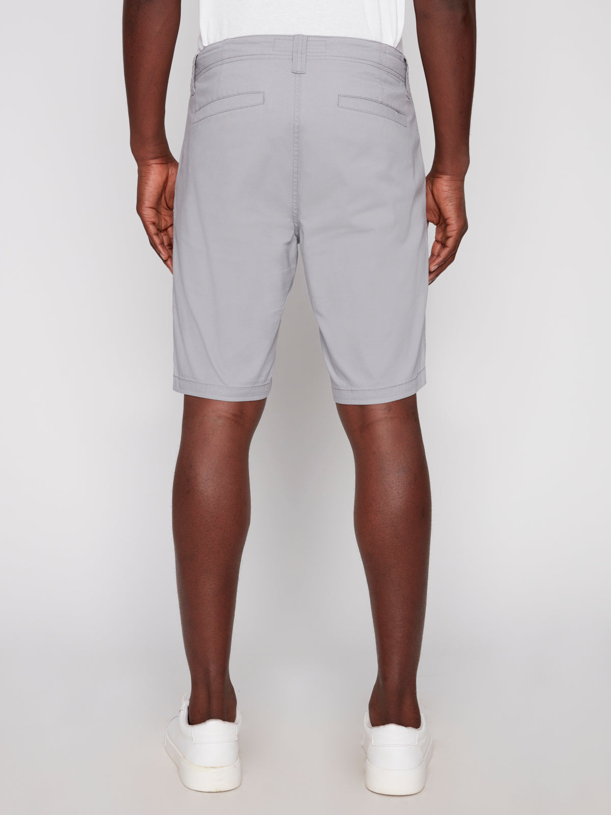 A person faces away, wearing Projek Raw GUSTAVE-ALLOY light gray knee-length shorts with two back welt pockets and visible stitching, paired with a white shirt and sneakers. The background is plain and light-colored.