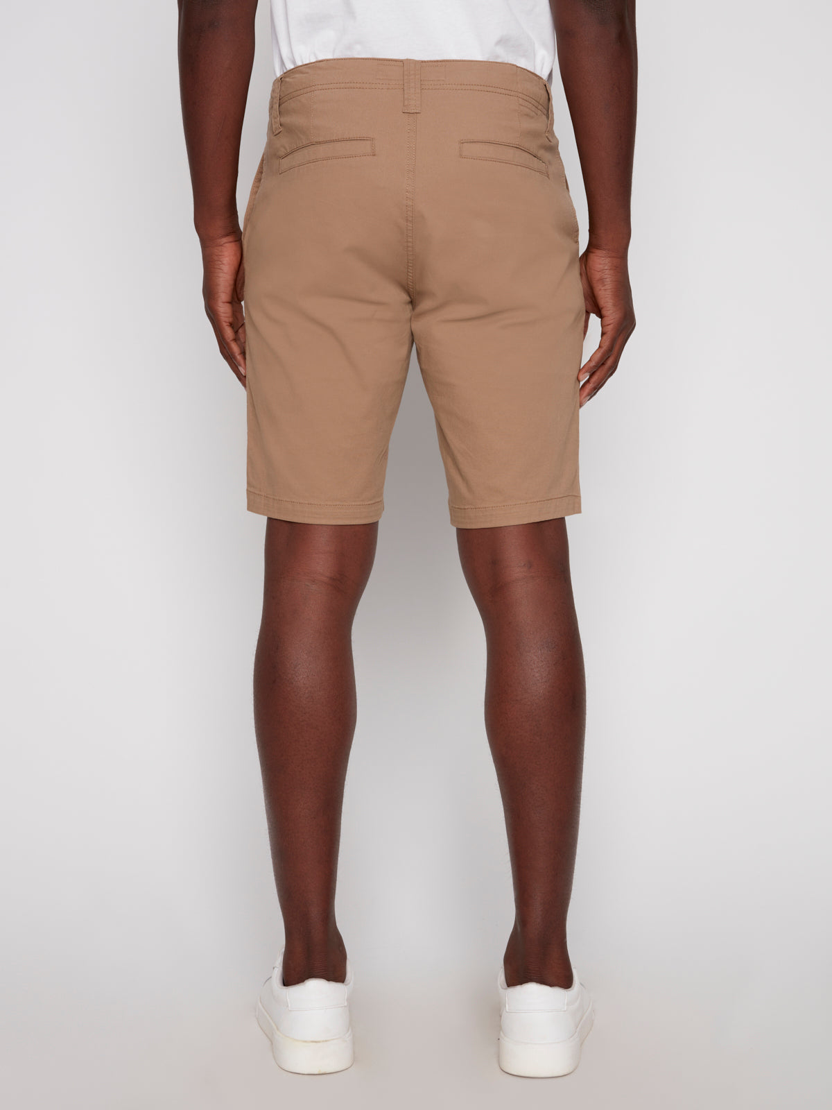 A person is seen from the back wearing Projek Raw’s GUSTAVE-TAN knee-length shorts, paired with a white shirt and white sneakers, standing against a plain white background.