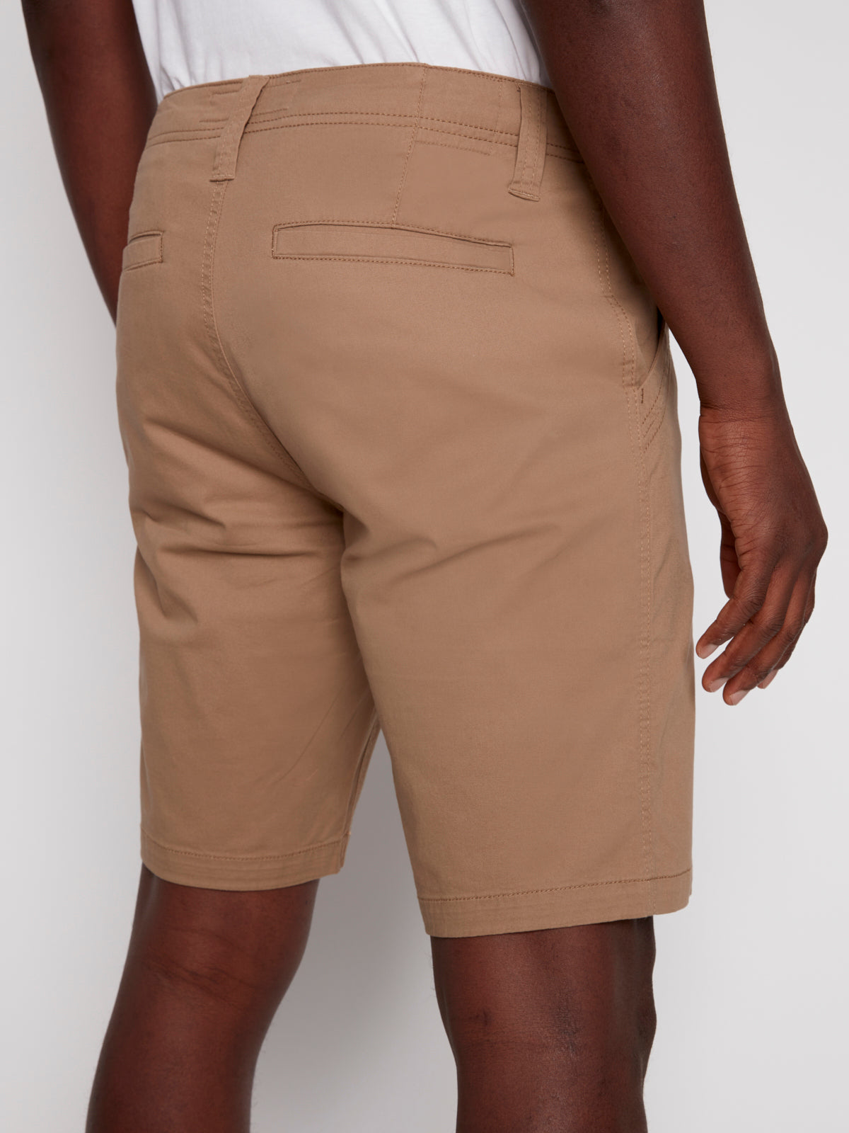 A person faces away, showcasing the back view of the Projek Raw GUSTAVE-TAN chino shorts with two welt pockets, paired with a white shirt.