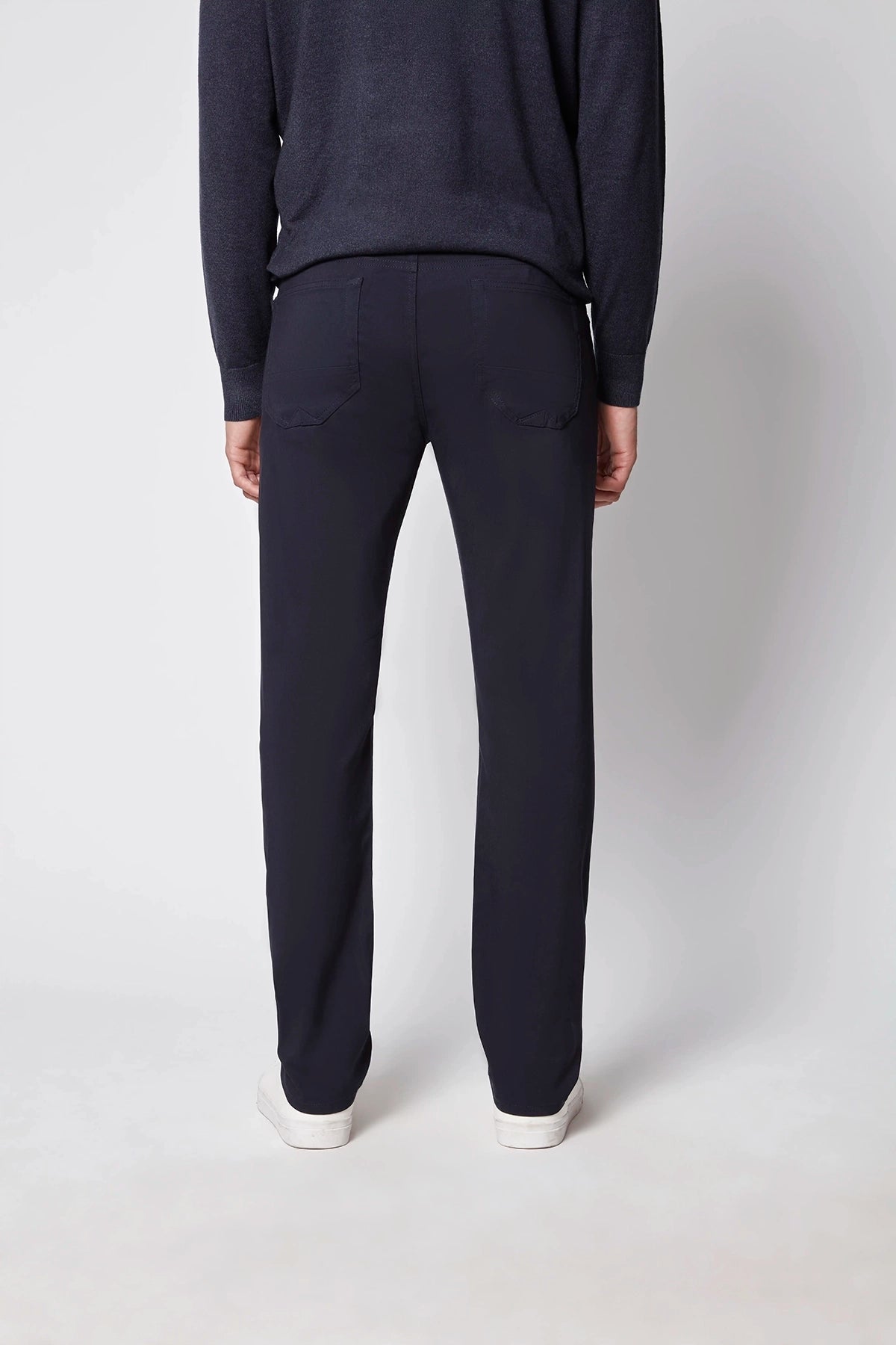Person stands with their back to the camera, wearing a dark long-sleeve top, PROJEK RAW Mens 5-pocket Classic Stretch Bungee Pants, and white sneakers against a light gray background.
