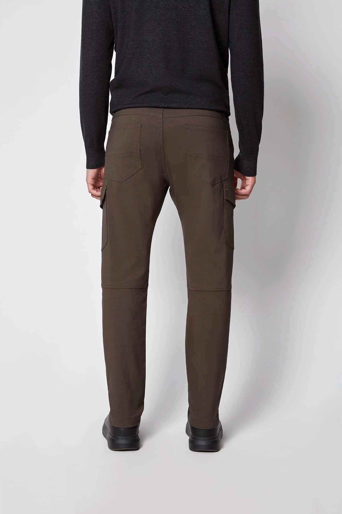 A person wearing PROJEK RAW Mens Stretch Bungee Cargo Pant in dark brown, paired with a black long-sleeve top and black shoes, stands facing away from the camera against a plain light gray background.